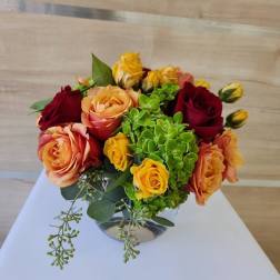 Low glass vase of orange, yellow, and red roses with bright green hydrangea