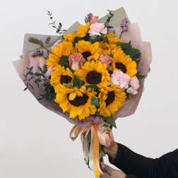 Handheld bouquet of sunflowers, pink roses, and carnations wrapped in pastel paper