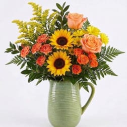 Bouquet of sunflowers, peach roses, and orange carnations in a green ceramic pitcher
