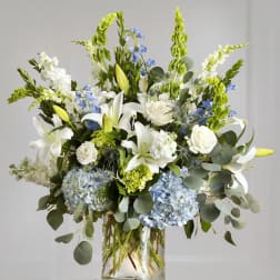 Tall bouquet of white lilies, roses, and blue hydrangeas in a glass vase