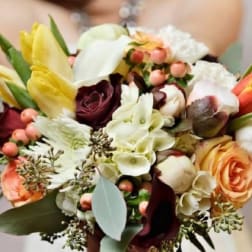 Hand-tied bouquet of ivory, peach, yellow, and burgundy flowers with berries, held by a person in white.