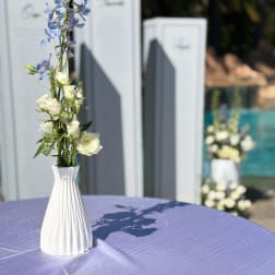 Tall white vase with white roses and blue flowers on a purple tablecloth