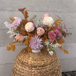 Pastel pink and peach floral arrangement with lavender mums and autumn leaves in a white vase on a woven basket.