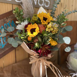 Rustic bouquet of yellow sunflowers, orange roses, and burgundy blooms wrapped in brown paper and raffia.