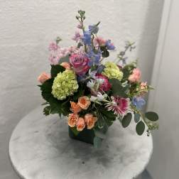 Pastel arrangement of pink and peach roses, green hydrangea and blue blooms in a green square vase