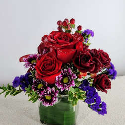Red roses and purple daisies arranged in a square glass vase