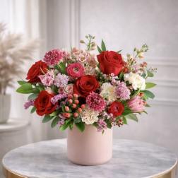 Mixed red and pink flower arrangement with roses and ranunculus in a pink vase on a round table.