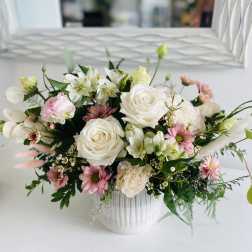 Low white ceramic vase filled with white roses, pink daisies, and soft pastel mixed flowers.