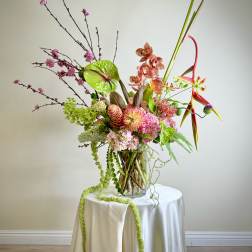 Tall modern arrangement with pink, green, and orange flowers in a glass vase on a draped round table