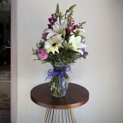 Tall mixed bouquet of white lilies and daisies with purple and pink flowers in a clear vase with a purple bow.