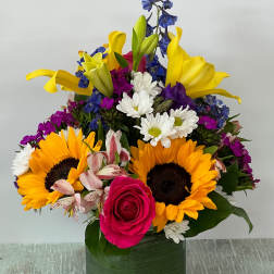 Colorful mixed bouquet in a square glass vase with sunflowers, lilies, roses, and daisies