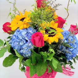 Tall mixed bouquet with yellow sunflowers, pink roses, and blue hydrangeas in a clear glass vase
