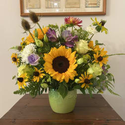 Mixed arrangement with sunflowers, yellow daisies, roses and purple blooms in a green ceramic vase