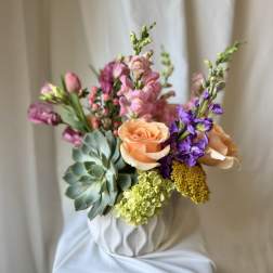 Bouquet of peach roses, purple blooms, and succulents in a white vase