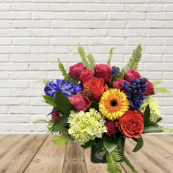 Mixed bouquet of roses, gerbera daisy, hydrangea, and blue blooms in a glass vase