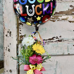 Tall vase of pink, yellow, and white gerbera daisies with a colorful round 'Good Luck' balloon.