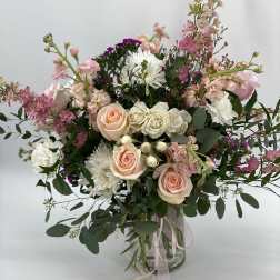Tall arrangement of peach roses, white mums, and pink blooms in a glass vase with trailing ribbons