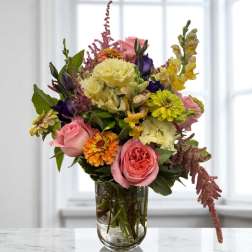 Mixed bouquet of pink roses, yellow carnations, and purple blooms in a glass vase