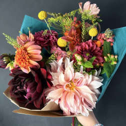 Handheld bouquet of pink, peach, and burgundy flowers wrapped in teal paper