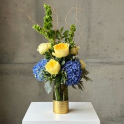 Yellow roses and blue hydrangeas in a gold vase with curly branches