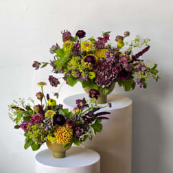 Two purple and yellow floral arrangements in gold vases on white pedestals