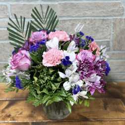 Round arrangement of pink roses, pink carnations, white alstroemeria, and purple blooms in a clear glass vase