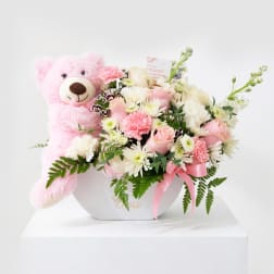 Pink teddy bear beside a pastel flower arrangement in a white box