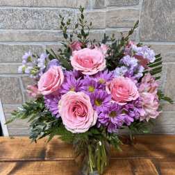 Pink roses and purple daisies arranged in a clear glass vase on a wooden surface.