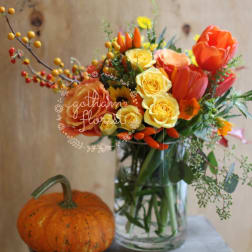 Orange and yellow flower bouquet in a clear glass vase with a pumpkin nearby