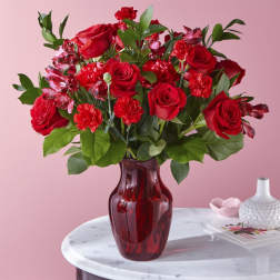 Arrangement of red roses and carnations in a red glass vase on a marble table.