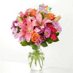 Mixed bouquet of pink lilies, orange roses, and purple mums in a clear glass vase with a Happy Birthday pick.