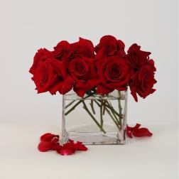 Red roses arranged in a clear square glass vase with scattered petals