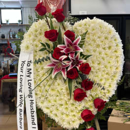 Heart-shaped white floral tribute with red roses and lilies, plus a memorial ribbon