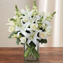 Arrangement of white lilies, roses, and snapdragons in a clear glass cylinder vase on a wooden table.