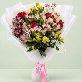 Mixed bouquet of pink, red, and yellow flowers wrapped in white paper with a pink ribbon