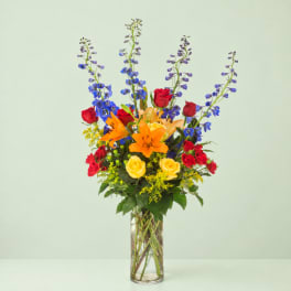 Tall mixed bouquet with red and yellow roses, orange lilies, and blue delphinium in a glass vase