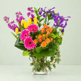 Colorful bouquet with pink gerberas, orange roses, yellow tulips, and purple irises in a clear vase