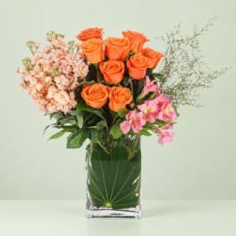Orange roses and pink flowers arranged in a clear square vase