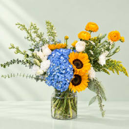 Mixed bouquet with blue hydrangea, sunflowers, white roses, and yellow ranunculus in a glass vase