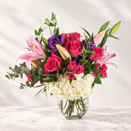 Bouquet of pink roses, lilies, and purple blooms in a clear glass vase