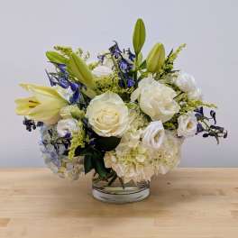 White roses, lilies, and hydrangeas with blue accents arranged in a low clear glass vase
