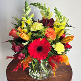 Mixed bouquet in a clear glass vase with red gerbera, roses, lilies, and yellow snapdragons.