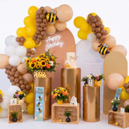 Birthday display with balloon arches, sunflowers, and a cake on gold pedestals