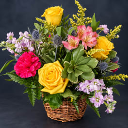 Mixed bouquet of yellow roses, pink carnations, and lavender flowers in a wicker basket