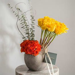 Yellow and orange roses arranged in a round gray vase with tall eucalyptus stems