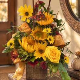 Yellow sunflowers and roses arranged in a rustic vase with ribbon and a butterfly pick.