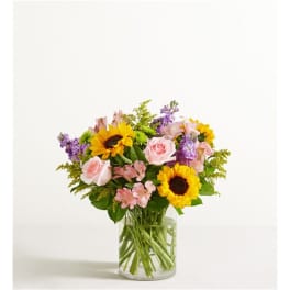 Mixed bouquet of sunflowers, pink roses, and purple flowers in a clear glass vase