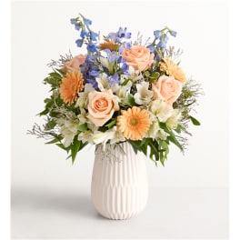 Mixed bouquet of peach roses, blue delphinium, and white flowers in a white vase