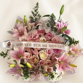 Pink lilies and roses arranged with a ribbon reading "Quieres ser mi novia?"