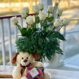 White roses in a glass vase with a teddy bear and gift box